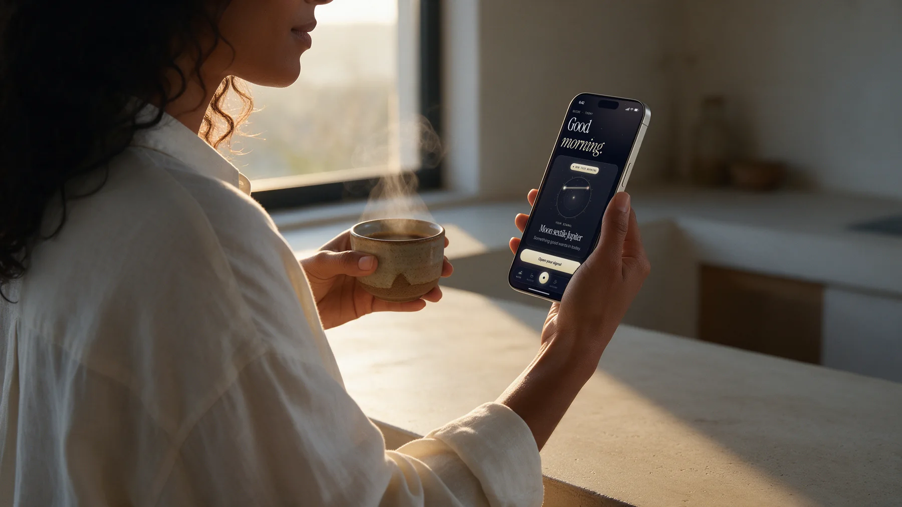 A woman at her kitchen bench at sunrise, coffee in one hand, reading her morning reading from Auriana on her phone